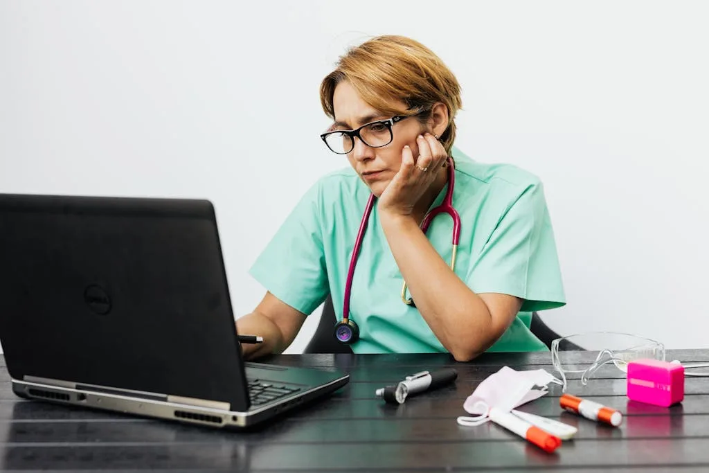 Focused nurse with eyeglasses working on a laptop surrounded by medical tools on a desk.