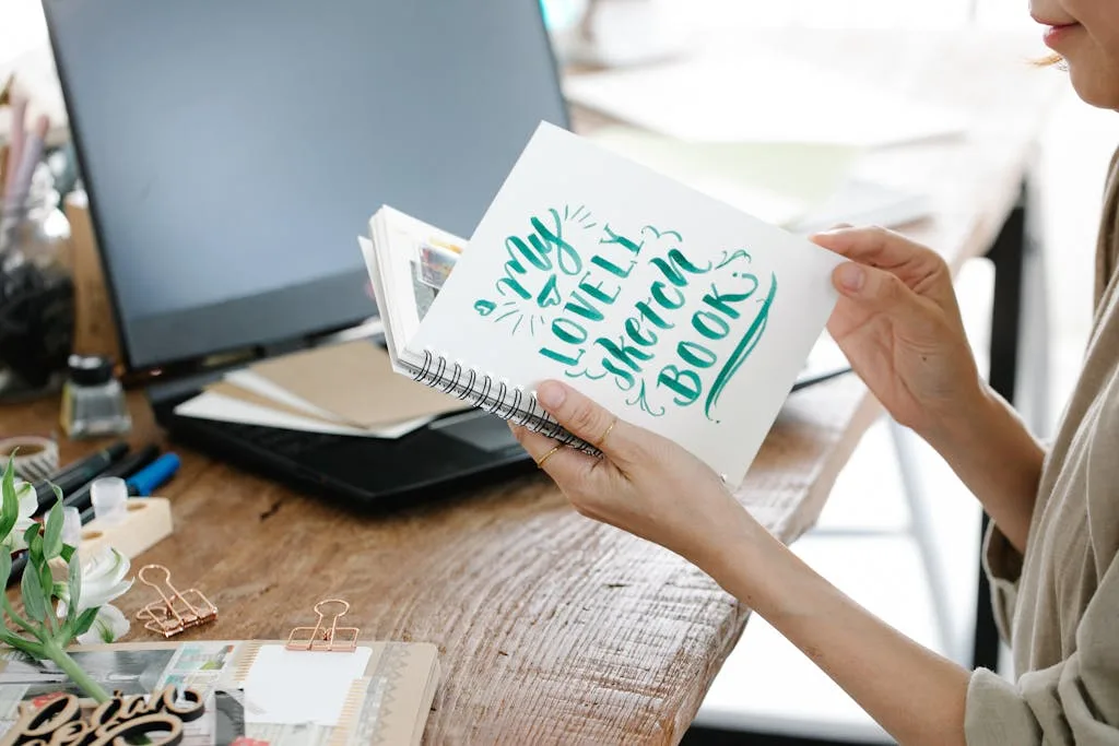 Woman holding sketchbook in a creative workspace with laptop, inspiring artistic environment.