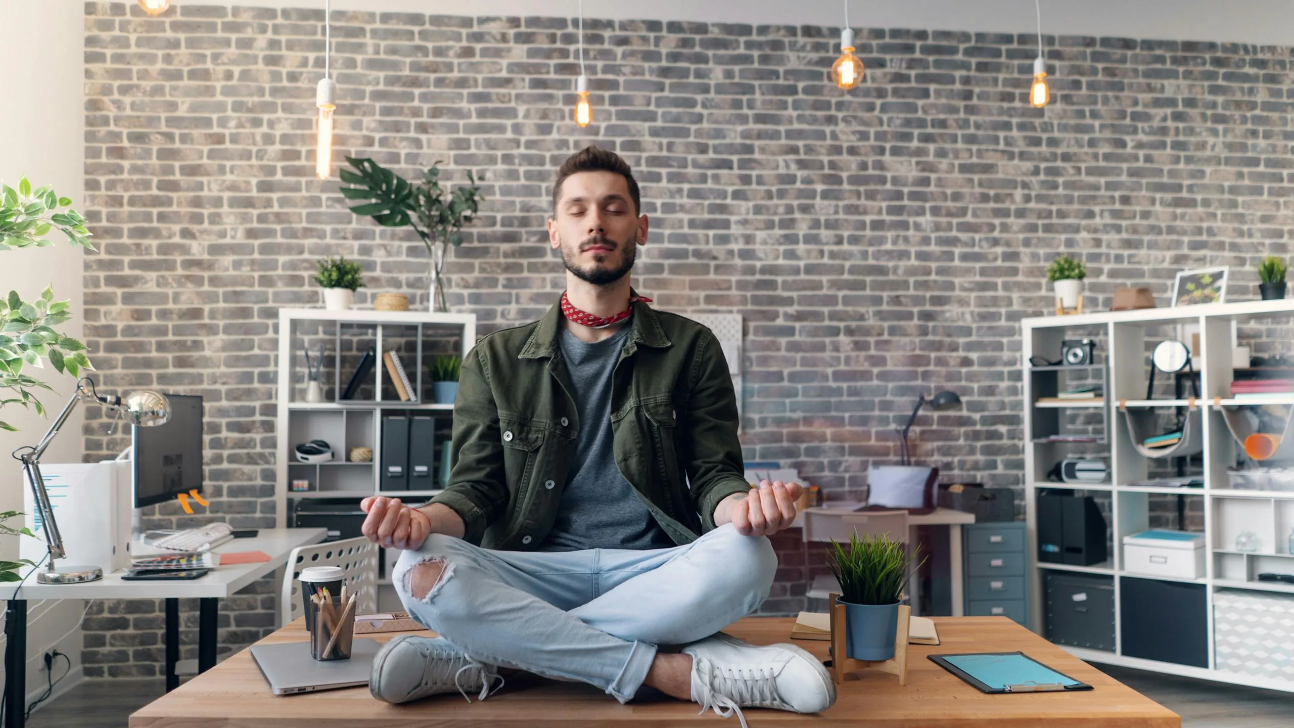 Young man practicing meditation on a desk in a modern office with brick walls.