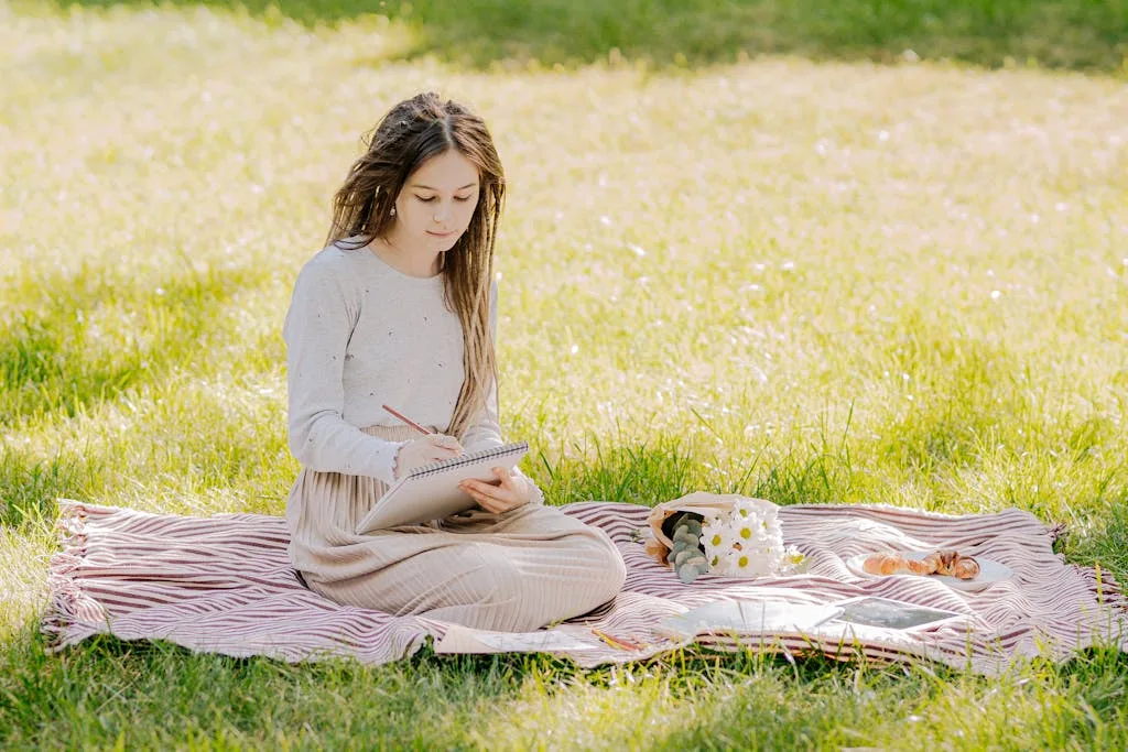 A young woman sitting on a blanket in a park, writing in a notebook on a sunny day.