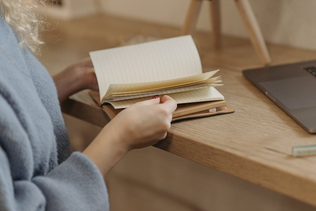 Person flipping through a notebook beside a laptop on a wooden table.