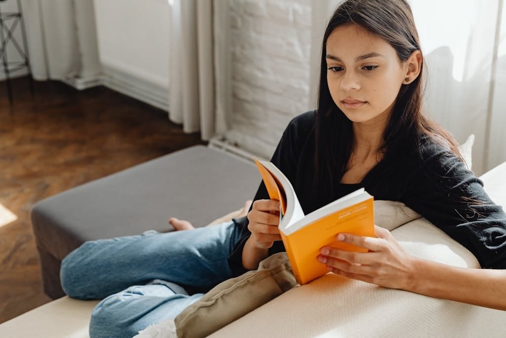 Young woman in black top and jeans reading a book indoors on a sofa.